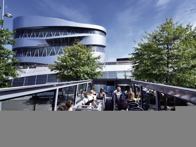Touristen sitzen in einem offenen Doppeldeckerbus vor dem modernen Mercedes-Benz Museum in Stuttgart, umgeben von Bäumen und blauem Himmel., © SMG, Pierre Polak Touristen sitzen in einem offenen Doppeldeckerbus vor dem modernen Mercedes-Benz Museum in Stuttgart, umgeben von Bäumen und blauem Himmel., © SMG, Pierre Polak