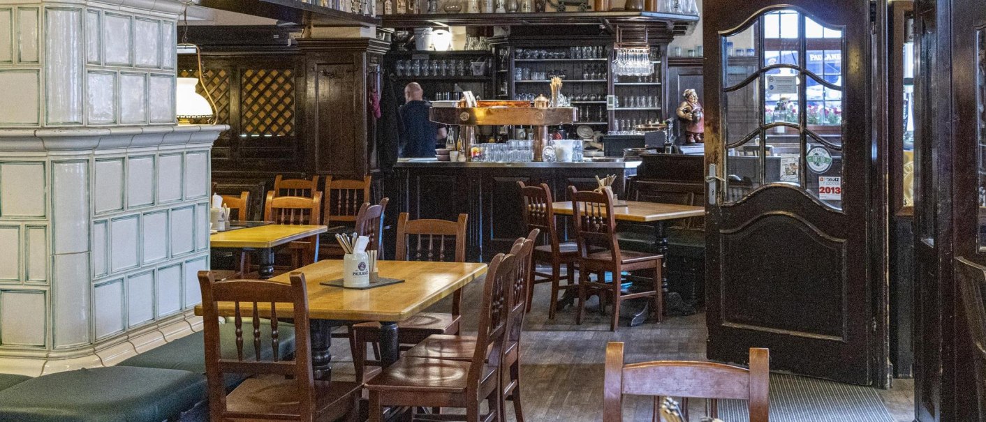Cozy interior of a restaurant with wooden tables and chairs, a bar in the background and a large tiled stove on the left., &copy; SMG Stuttgart Marketing GmbH - Sarah Schmid
