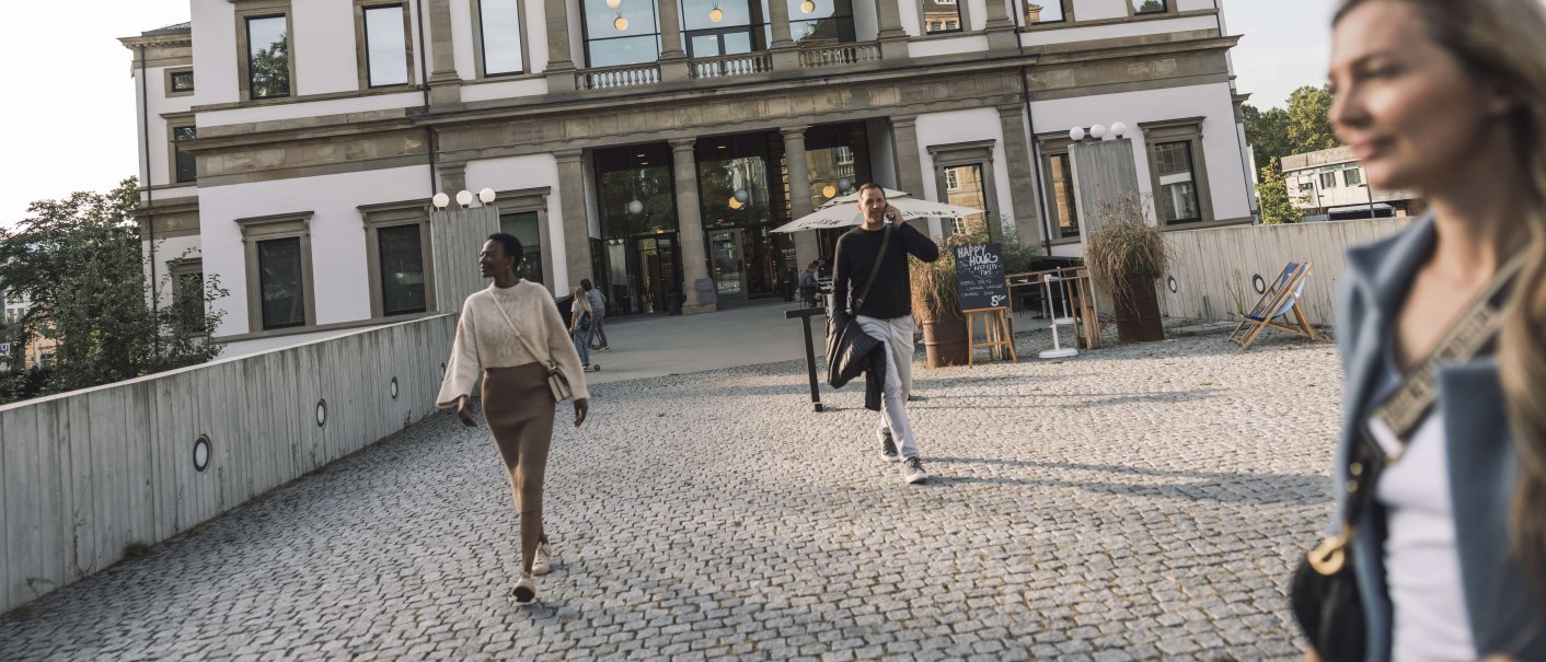 Das StadtPalais Stuttgart mit klassischer Fassade, Menschen gehen auf einem gepflasterten Weg davor. Im Hintergrund sind Bäume und ein sonniger Himmel., © Stuttgart-Marketing GmbH, WP Steinheisser
