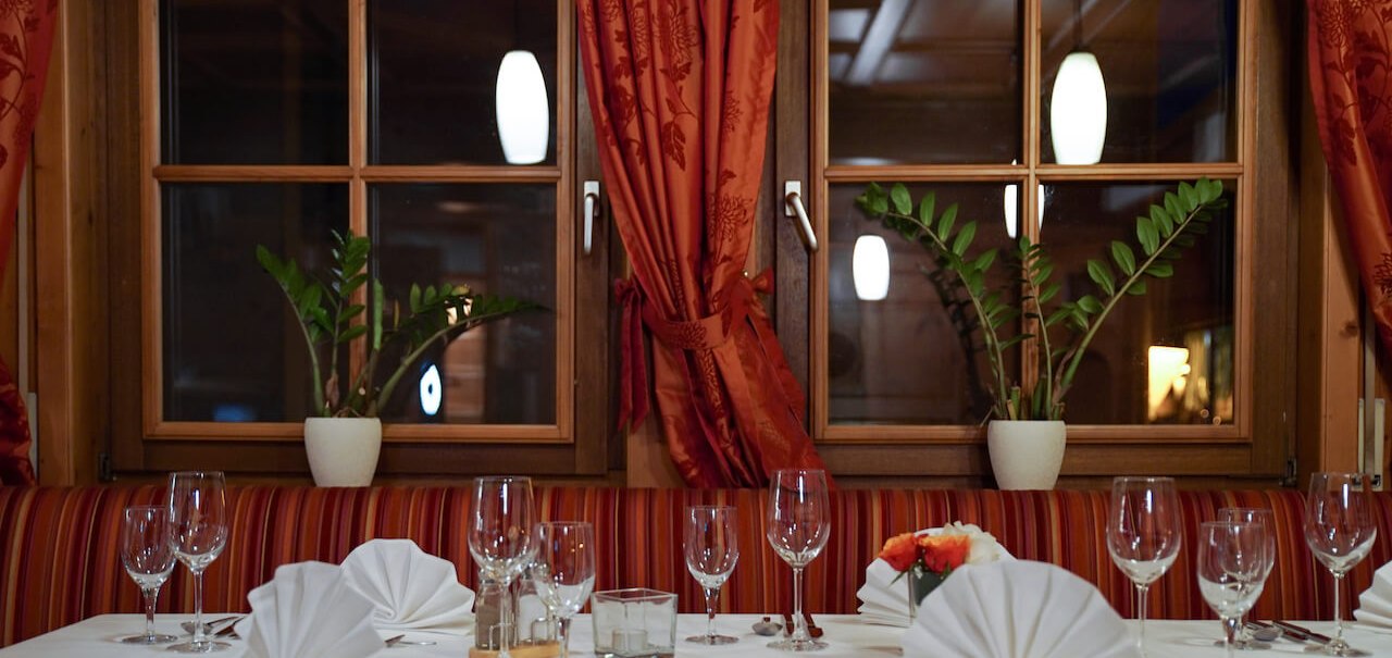 Ein stilvoll gedeckter Tisch in einem Restaurant mit wei&szlig;en Tischdecken, gefalteten Servietten und Weingl&auml;sern. Im Hintergrund sind Fenster mit roten Vorh&auml;ngen., &copy; Hetzel Hotel Stuttgart