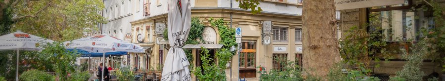A bistro on Wilhelmsplatz with empty tables and chairs outside, surrounded by green plants and trees., &copy; Martina Denker