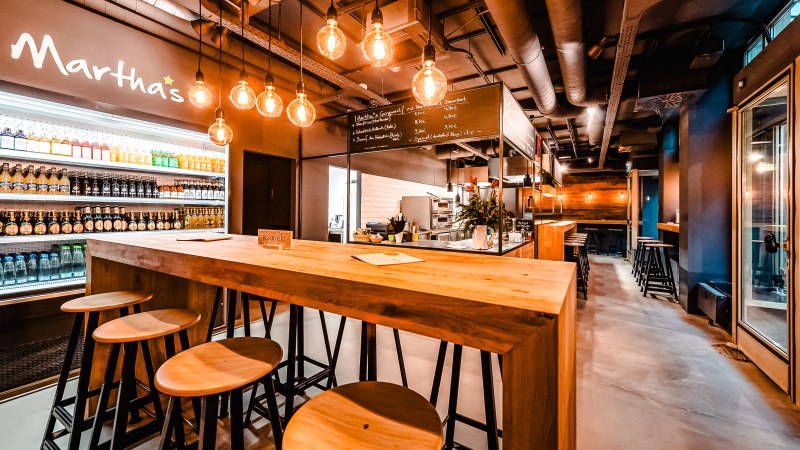 Modernes Café mit Holztischen, Hockern und hängenden Glühbirnen. Getränkekühlschrank und Menütafel im Hintergrund. Gemütliche Atmosphäre., © Andreas Dalferth Modernes Café mit Holztischen, Hockern und hängenden Glühbirnen. Getränkekühlschrank und Menütafel im Hintergrund. Gemütliche Atmosphäre., © Andreas Dalferth