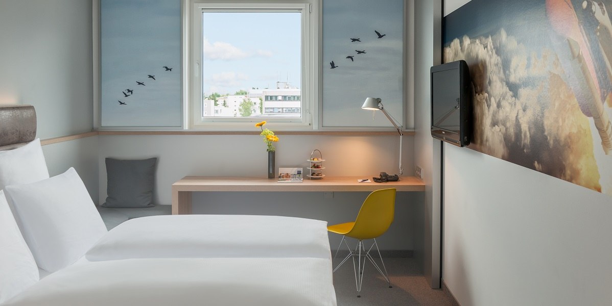 Modernes Doppelzimmer mit wei&szlig;em Bett, Schreibtisch, gelbem Stuhl und Fenster mit Stadtblick. Wanddekoration mit Vogelmotiv., &copy; Essential by Dorint