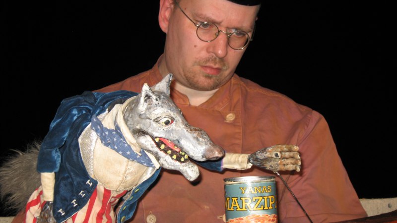 Ein Mann mit Brille und M&uuml;tze h&auml;lt eine Wolfspuppe, die nach einer Dose Marzipan greift. Die Szene wirkt humorvoll und skurril., &copy; Figurentheater Oliver K&ouml;hler