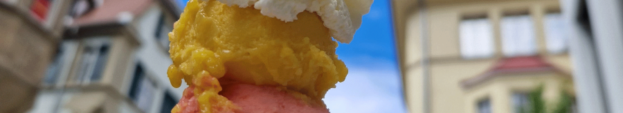 An ice cream cone with three scoops in different colors and whipped cream, against a background of buildings and blue sky., &copy; kesselglueck.de