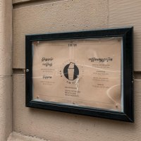 Schild der TinTin Bar mit Öffnungszeiten und Preisen an einer Wand neben einer weißen Tür., © SMG, Sarah Schmid
