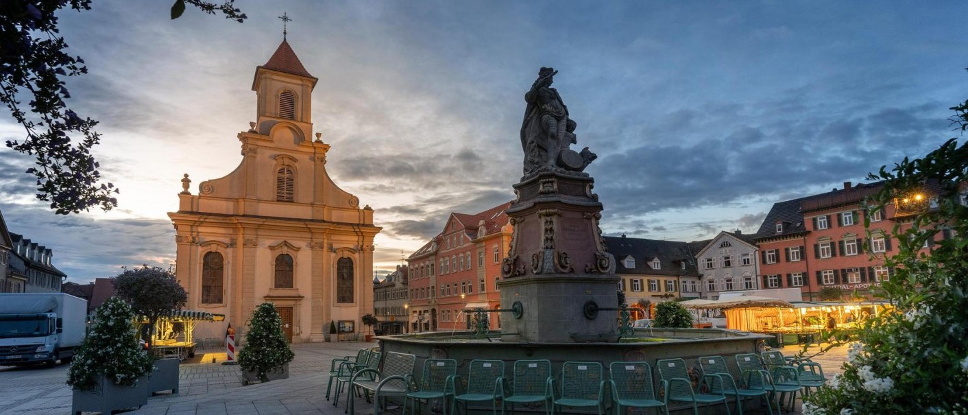 Evening scene on Ludwigsburg's market square, featuring the illuminated church and fountain. Chairs are arranged around the fountain, with market stalls in the background., © © Stuttgart-Marketing GmbH, Martina Denker Evening scene on Ludwigsburg's market square, featuring the illuminated church and fountain. Chairs are arranged around the fountain, with market stalls in the background., © © Stuttgart-Marketing GmbH, Martina Denker
