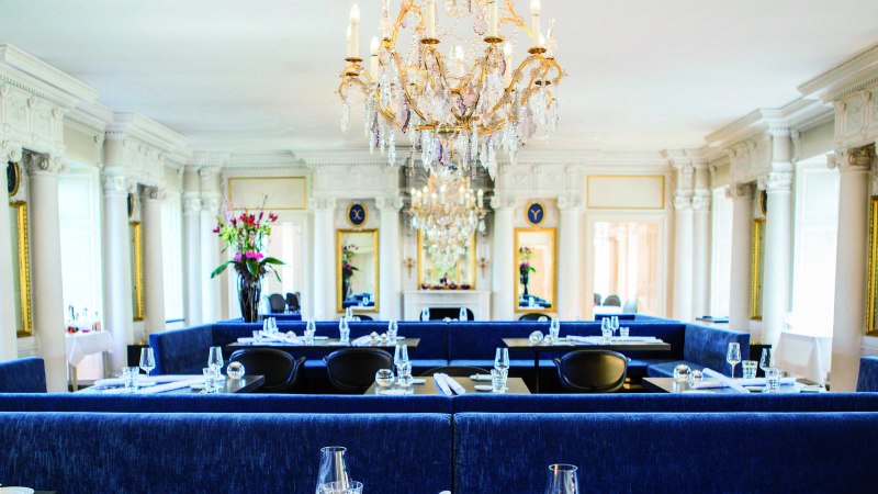 Elegant restaurant with chandelier, blue benches and white pillars. Flower arrangements and laid tables create a luxurious atmosphere., © Speisemeisterei Elegant restaurant with chandelier, blue benches and white pillars. Flower arrangements and laid tables create a luxurious atmosphere., © Speisemeisterei