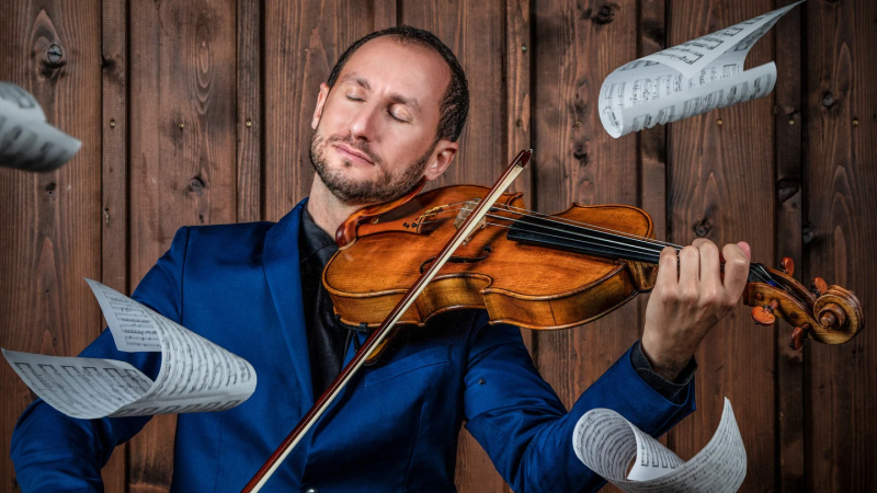 A violinist in a blue suit plays the violin passionately, surrounded by floating sheet music against a wooden background., &copy; privat