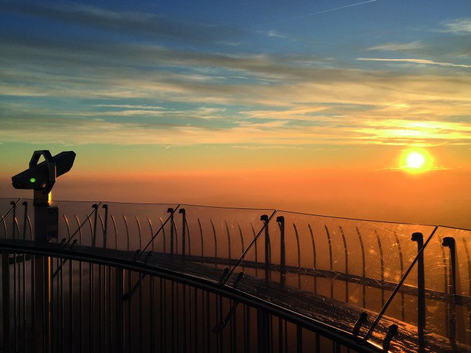 The observation deck of a TV tower at sunrise. A telescope and a glass railing are visible as the sun rises on the horizon., © SWR Media Services GmbH / Fernsehturm Stuttgart The observation deck of a TV tower at sunrise. A telescope and a glass railing are visible as the sun rises on the horizon., © SWR Media Services GmbH / Fernsehturm Stuttgart