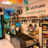 Ein Feinkostladen mit lateinamerikanischen Produkten, bunten Dekorationen und einer Kasse. Regale sind mit Snacks und Getränken gefüllt., © Feinkost Latino Ventura Ein Feinkostladen mit lateinamerikanischen Produkten, bunten Dekorationen und einer Kasse. Regale sind mit Snacks und Getränken gefüllt., © Feinkost Latino Ventura