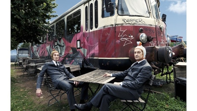Zwei M&auml;nner in Anz&uuml;gen sitzen an einem Tisch vor einem bunt bemalten, alten Zug. Der Himmel ist blau, und die Umgebung wirkt l&auml;ndlich., &copy; Rosenau Kultur e.V.