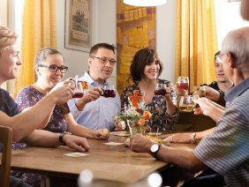 Eine Gruppe von Menschen sitzt an einem Tisch und stößt mit Getränken an. Sie wirken fröhlich und entspannt. Im Hintergrund sind gelbe Vorhänge., © Stuttgart-Marketing GmbH / Winkler