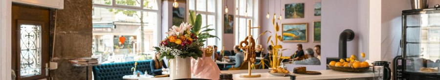 Gem&uuml;tliches Caf&eacute; mit Blumen und Brezeln auf der Theke, blauen Sofas und Kunst an den W&auml;nden. Gro&szlig;e Fenster lassen viel Licht herein., &copy; SMG, Sarah Schmid