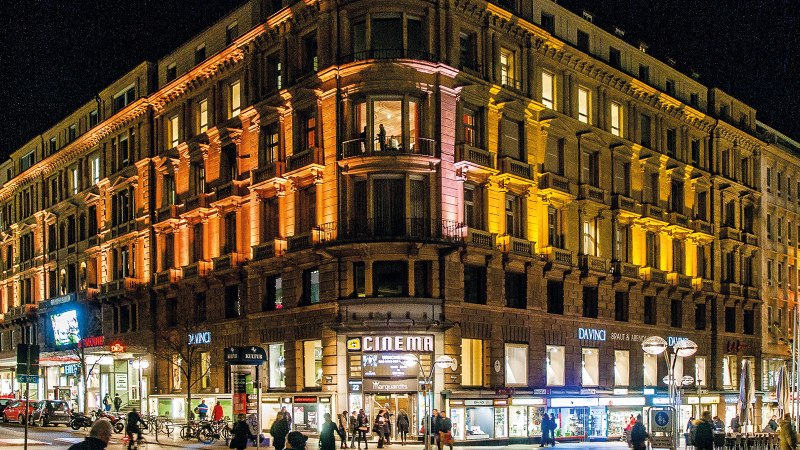 Ein großes, beleuchtetes Gebäude bei Nacht mit Geschäften im Erdgeschoss und Menschen auf der Straße., © Schauspielbühnen in Stuttgart