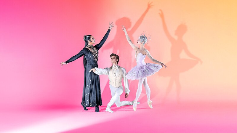 Drei Balletttänzer in eleganten Kostümen posieren vor einem farbigen Hintergrund. Zwei Tänzer stehen, während einer kniet. Schatten sind sichtbar., © Württembergische Staatstheater Stuttgart Drei Balletttänzer in eleganten Kostümen posieren vor einem farbigen Hintergrund. Zwei Tänzer stehen, während einer kniet. Schatten sind sichtbar., © Württembergische Staatstheater Stuttgart