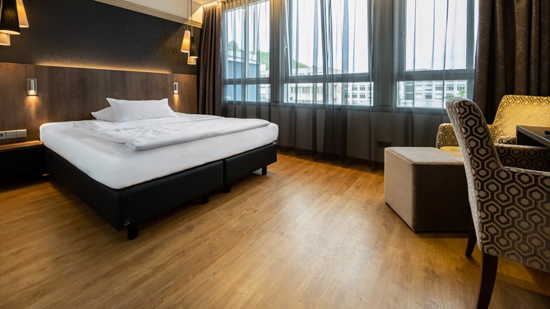 Modernes Hotelzimmer mit großem Bett, Holzboden, stilvollen Lampen und gemütlichen Sesseln vor großen Fenstern., © Kronenhotel Stuttgart