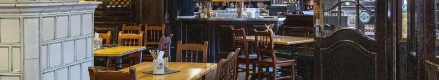 Cozy interior of a restaurant with wooden tables and chairs, a bar in the background and a large tiled stove on the left., © SMG Stuttgart Marketing GmbH - Sarah Schmid