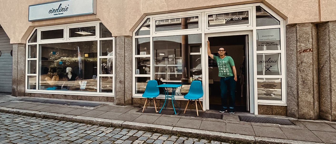 Modegeschäft 'einelinie' mit Schaufenster und Eingang. Ein Mann steht in der Tür, blaue Stühle und Tisch davor auf Kopfsteinpflaster., © StuttgARTfactory