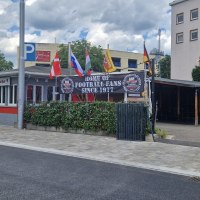 Bar mit Fahnen und Schild "Home of Football Fans Since 1977". Gebäude mit roten und grauen Akzenten, umgeben von Bäumen und einem Parkplatz., © Stuttgart-Marketing GmbH