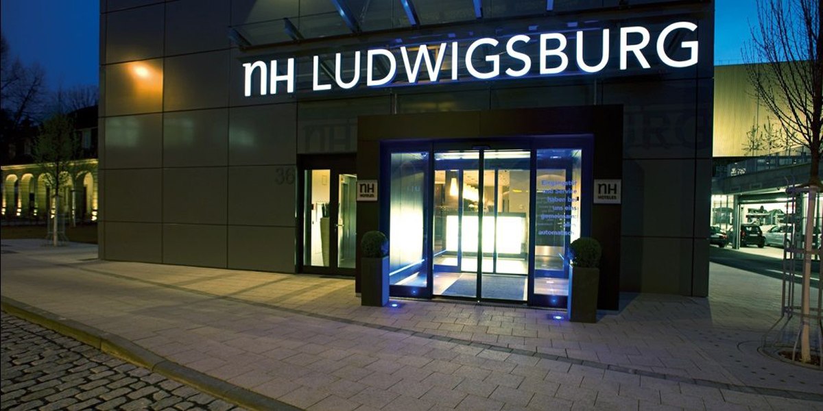 The entrance to the NH Hotel in Ludwigsburg at night, illuminated by blue light. The "NH LUDWIGSBURG" sign is clearly visible., © NH Ludwigsburg The entrance to the NH Hotel in Ludwigsburg at night, illuminated by blue light. The "NH LUDWIGSBURG" sign is clearly visible., © NH Ludwigsburg