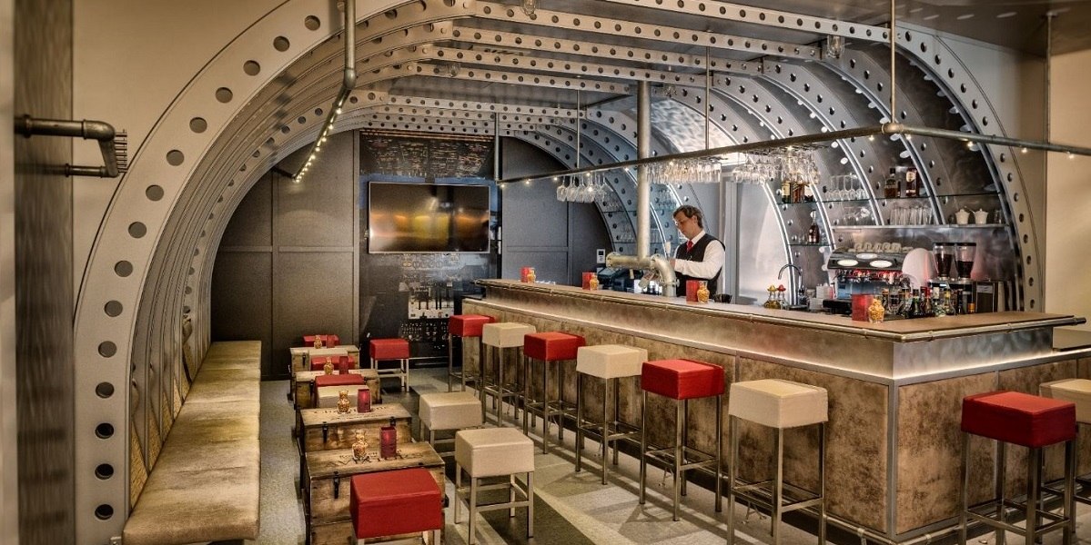 Moderne Bar mit industriellem Design, Metallb&ouml;gen und Bartresen. Rote und wei&szlig;e Hocker, gem&uuml;tliche Sitzbereiche und ein Barkeeper hinter der Theke., &copy; Essential by Dorint