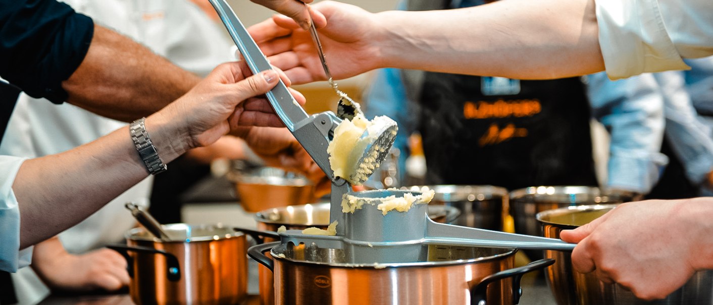 Mehrere Personen drücken Kartoffeln durch eine Presse in einen Topf. Im Hintergrund sind weitere Töpfe und eine Person mit Schürze zu sehen., © ILZHÖFER´s Event-Kochschule Mehrere Personen drücken Kartoffeln durch eine Presse in einen Topf. Im Hintergrund sind weitere Töpfe und eine Person mit Schürze zu sehen., © ILZHÖFER´s Event-Kochschule