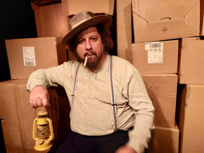 Ein Mann mit Hut und Zigarette sitzt vor gestapelten Kartons und hält eine gelbe Laterne in der Hand., © Theater am Olgaeck Ein Mann mit Hut und Zigarette sitzt vor gestapelten Kartons und hält eine gelbe Laterne in der Hand., © Theater am Olgaeck