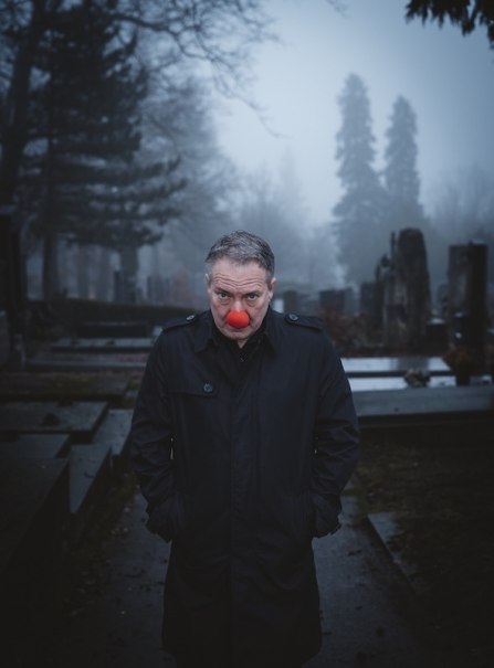 A person with a red clown's nose is standing in a foggy cemetery. She is wearing a dark coat and has her hands in her pockets., &copy; Renitenztheater Stuttgart e.V.