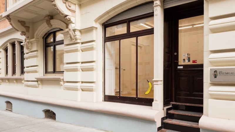 Eingang einer Galerie mit dem Schild 'Die Raumgalerie'. Eine Banane ist an der Glastür angebracht. Das Gebäude hat eine verzierte Fassade., © Dirk Wilhelmy Fotografie