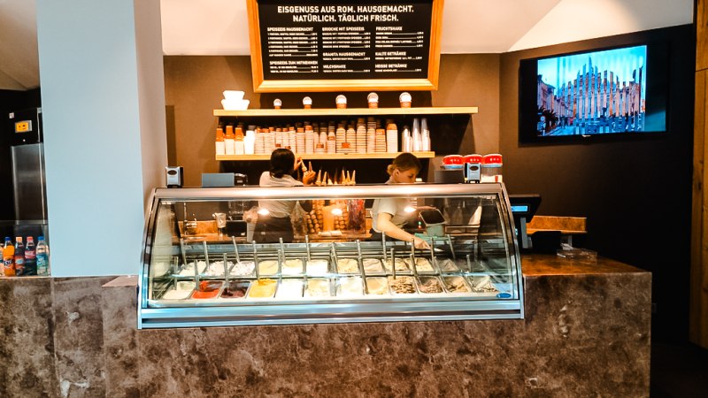 Eisdiele mit Theke, gefüllt mit verschiedenen Eissorten. Zwei Personen arbeiten dahinter. An der Wand ein Menü und ein Bildschirm., © Old Bridge Gelateria Stuttgart