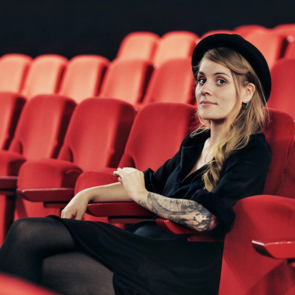 Eine Frau mit Hut und tätowiertem Arm sitzt entspannt in einem leeren Kino auf roten Sitzen., © Renitenztheater Stuttgart e.V. Eine Frau mit Hut und tätowiertem Arm sitzt entspannt in einem leeren Kino auf roten Sitzen., © Renitenztheater Stuttgart e.V.