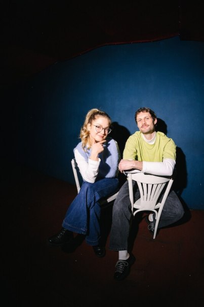 Zwei Personen sitzen l&auml;ssig auf wei&szlig;en St&uuml;hlen vor einer blauen Wand. Die Frau tr&auml;gt eine Brille und l&auml;chelt, der Mann tr&auml;gt ein gr&uuml;nes T-Shirt., &copy; Rosenau Kultur e.V.