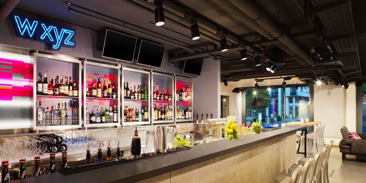 Moderne Bar mit beleuchtetem Spirituosenregal, Tresen mit Gläsern und Früchten. Neonschild 'W XYZ' an der Wand, stilvolle Beleuchtung., © Aloft Stuttgart