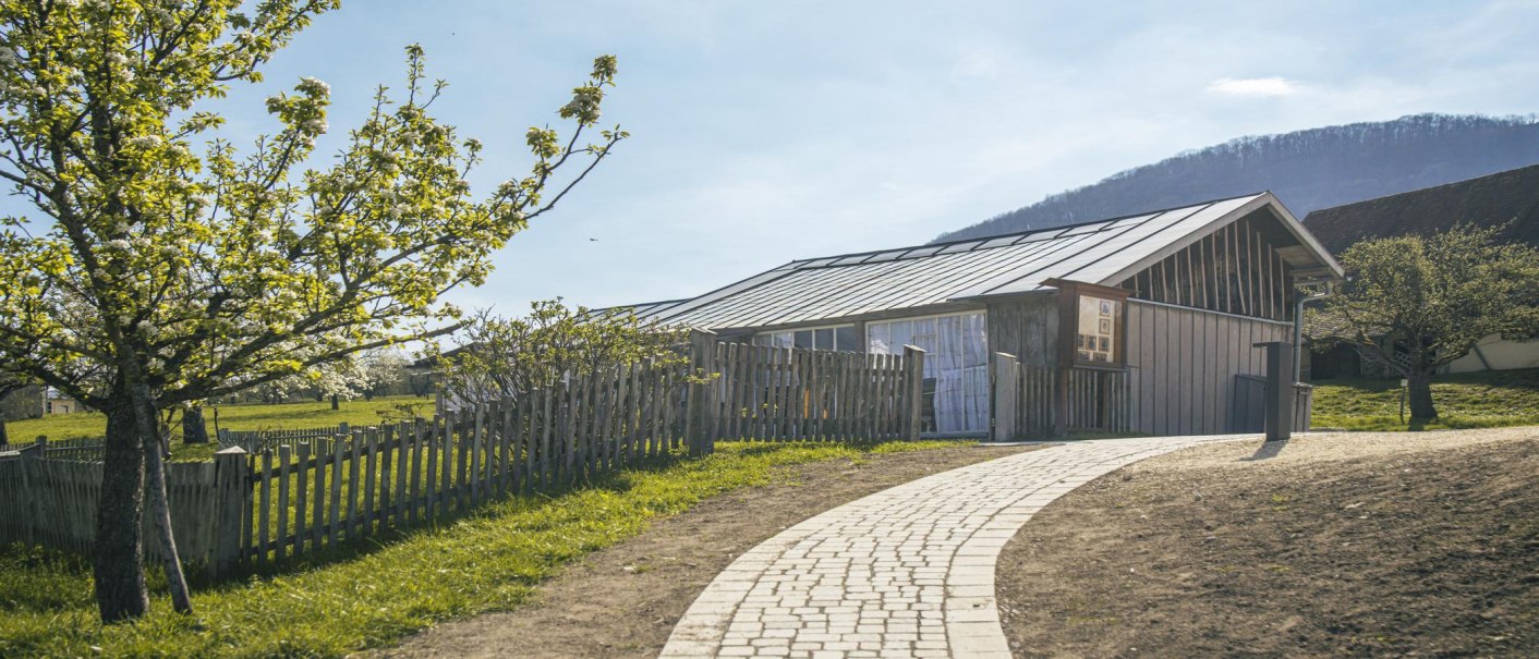 A modern building in the Beuren open-air museum, surrounded by blossoming trees and a wooden fence. A paved path leads to the entrance., © SMG Stuttgart Marketing GmbH - Sarah Schmid A modern building in the Beuren open-air museum, surrounded by blossoming trees and a wooden fence. A paved path leads to the entrance., © SMG Stuttgart Marketing GmbH - Sarah Schmid