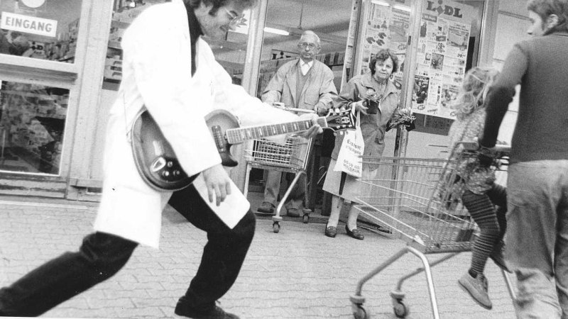 Ein Mann in einem wei&szlig;en Mantel spielt Gitarre vor einem Supermarkt. Passanten mit Einkaufswagen beobachten die Szene interessiert., &copy; Rock&rsquo;n&rsquo;Roll Diktator