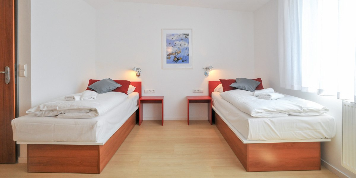 A bright twin room with two single beds, red nightstands, and wall sconces. A picture hangs on the white wall; the window has white curtains., &copy; G&auml;stehaus ABZ Stuttgart