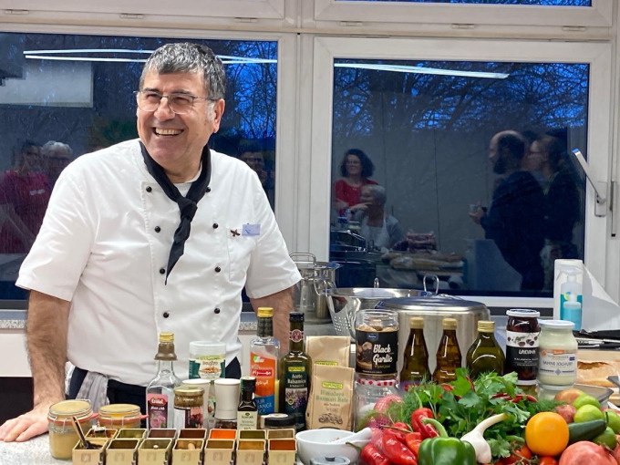 A chef in a white uniform presents a selection of ingredients and spices in a kitchen. People can be seen in the background., © Kultweine-Kochwelten-Kunstücke - Kochschule Sonja Lenz A chef in a white uniform presents a selection of ingredients and spices in a kitchen. People can be seen in the background., © Kultweine-Kochwelten-Kunstücke - Kochschule Sonja Lenz