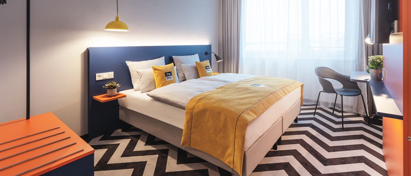 A modern hotel room with a double bed, a yellow comforter, a blue headboard, a zigzag rug, and a desk with a chair., © the niu Form - Novum Hospitality A modern hotel room with a double bed, a yellow comforter, a blue headboard, a zigzag rug, and a desk with a chair., © the niu Form - Novum Hospitality