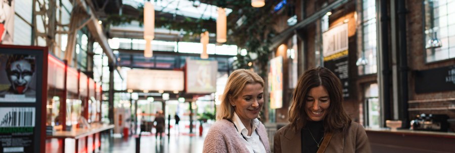 Two women are standing in the foyer of a theater looking at tickets. Posters and modern architecture can be seen in the background., © Stuttgart-Marketing GmbH, wpsteinheisser