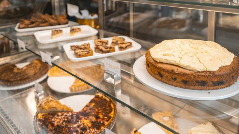 Eine Kuchenvitrine in einem Café zeigt verschiedene Kuchen und Gebäckstücke, darunter ein großer Kuchen mit heller Glasur und kleine Gebäckstücke auf Tabletts., © SMG, Sarah Schmid