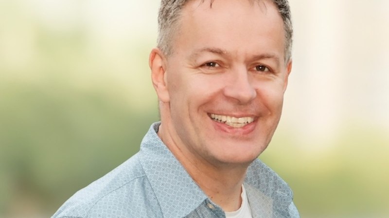 A man with gray hair and a blue shirt smiles at the camera. The background is blurred and greenish., &copy; Renitenztheater Stuttgart e.V.