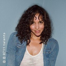 Eine Frau mit lockigem Haar trägt eine Jeansjacke und ein weißes Oberteil vor einem blauen Hintergrund., © links im Bild