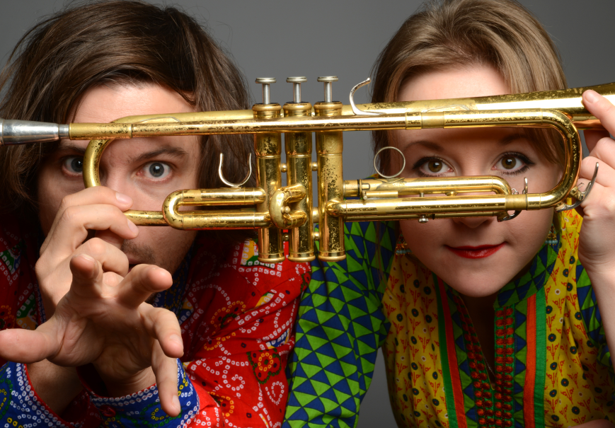 Zwei Personen in bunten Hemden blicken durch eine Trompete. Sie halten das Instrument vor ihre Gesichter und schauen neugierig hindurch., © Matria Zwei Personen in bunten Hemden blicken durch eine Trompete. Sie halten das Instrument vor ihre Gesichter und schauen neugierig hindurch., © Matria