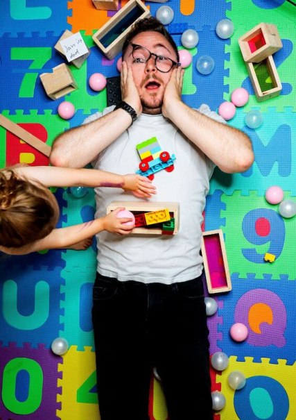 Ein Mann liegt &uuml;berrascht auf einer bunten Spielmatte, umgeben von Baukl&ouml;tzen und B&auml;llen. Ein Kind reicht ihm Spielzeug., &copy; Theaterhaus Stuttgart e.V.