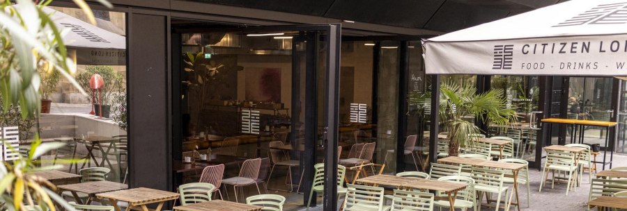 Outdoor area of a caf&eacute; with wooden tables and green chairs. An awning bears the lettering 'Citizen Long'. Plants decorate the area., &copy; SMG, Sarah Schmid