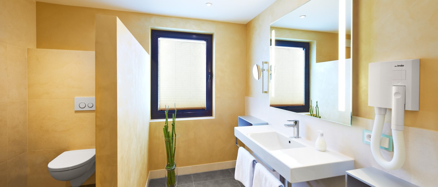 Modernes Badezimmer mit gelben Wänden, großem Spiegel, Waschbecken, Handtüchern und einer Toilette in einer Nische., © Hirsch Hotel