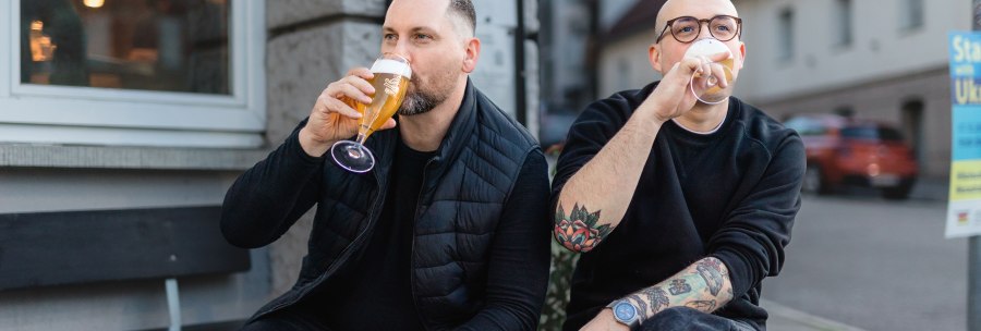 Two men are sitting on a wooden pallet in front of a building drinking beer. One of them wears glasses and has tattooed arms., © © Jaimee Moses All Rights Reserved Two men are sitting on a wooden pallet in front of a building drinking beer. One of them wears glasses and has tattooed arms., © © Jaimee Moses All Rights Reserved