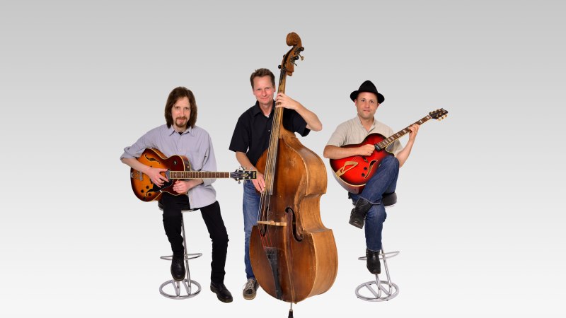 Ein Jazztrio mit zwei Gitarristen und einem Kontrabassisten posiert vor einem grauen Hintergrund. Alle sitzen auf Hockern., &copy; Saite16