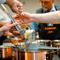 Mehrere Personen drücken Kartoffeln durch eine Presse in einen Topf. Im Hintergrund sind weitere Töpfe und eine Person mit Schürze zu sehen., © ILZHÖFER´s Event-Kochschule Mehrere Personen drücken Kartoffeln durch eine Presse in einen Topf. Im Hintergrund sind weitere Töpfe und eine Person mit Schürze zu sehen., © ILZHÖFER´s Event-Kochschule